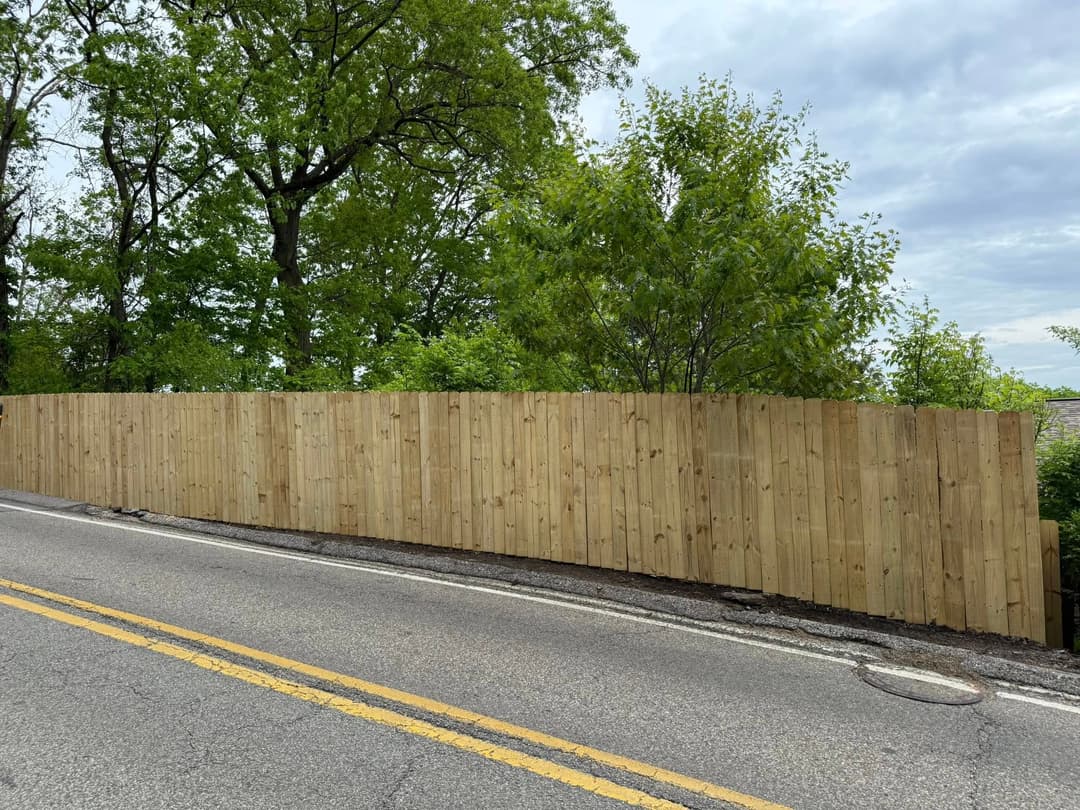 wooden privacy fence along a roadside with greenery in the background and cloudy sky