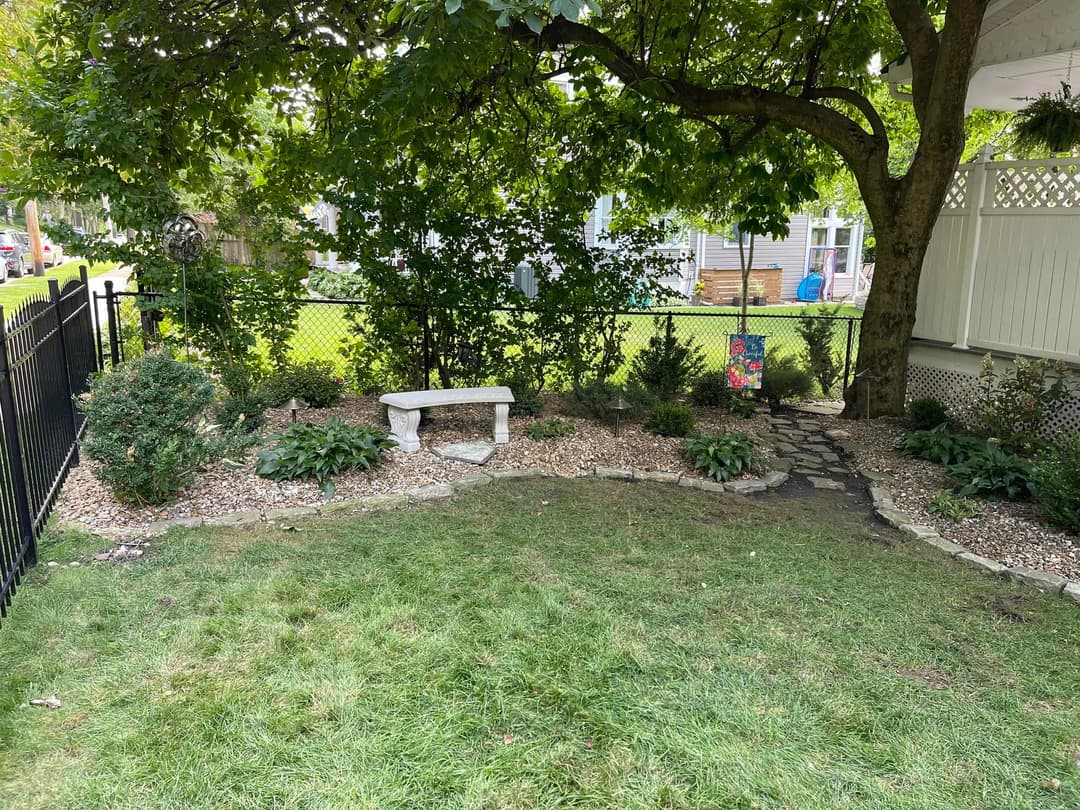 Cozy garden area with stone bench, lush plants, and bordered by a black fence.