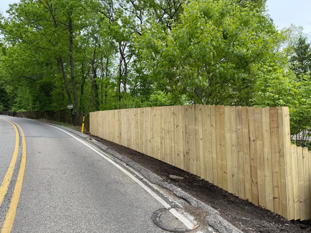 New wooden fence along a roadside, surrounded by lush green trees and foliage.