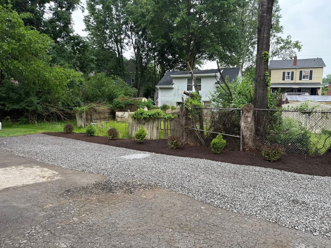 Gravel pathway and landscaped garden with shrubs beside a fenced yard and trees.